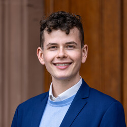 Felix Haas hat braunen Haare. Er trägt ein weiß Hemd und darüber einen hellblauen Pullover und ein dunkelblaues Jackett.