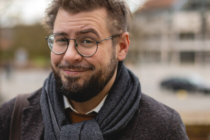 Hiram Kümper. Hiram Kümper trägt Brille, einen blauen Schal und ein schwarzes Sakko. Über seiner Schulter hängt eine braune Tasche.