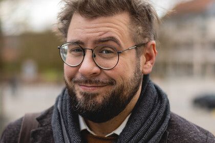 Hiram Kümper. Hiram Kümper trägt Brille, einen blauen Schal und ein schwarzes Sakko. Über seiner Schulter hängt eine braune Tasche.