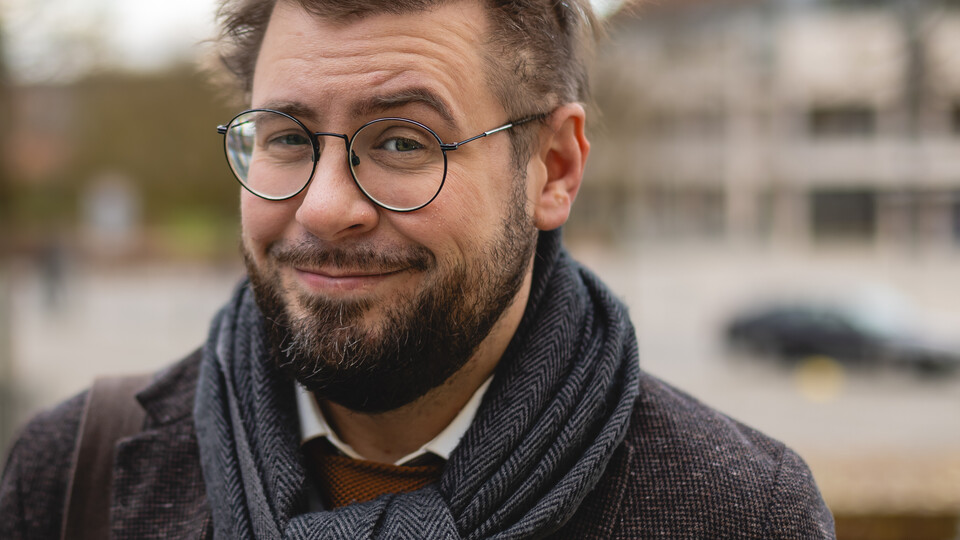 Hiram Kümper. Hiram Kümper trägt Brille, einen blauen Schal und ein schwarzes Sakko. Über seiner Schulter hängt eine braune Tasche.