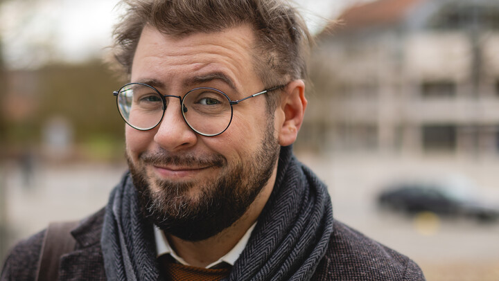 Hiram Kümper. Hiram Kümper trägt Brille, einen blauen Schal und ein schwarzes Sakko. Über seiner Schulter hängt eine braune Tasche.