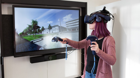 Eine Person trägt eine VR-Brille und zwei Controller in ihren Händen. Auf einem Bildschirm hinter ihr ist ein Teil des Campus der Uni Mannheim zu sehen.