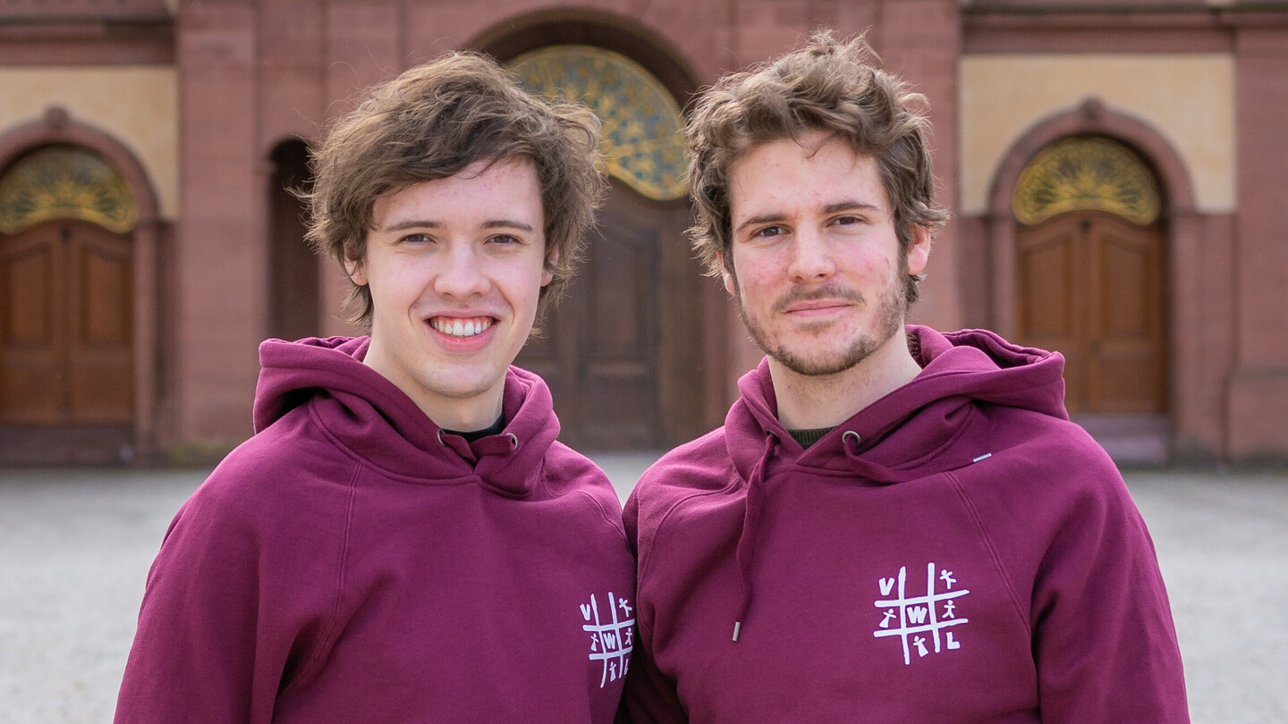 Zwei Studierende vor dem Mannheimer Schloss. Sie tragen beide kurze, braune Haare und einen Hoodie der Fachschaft VWL.