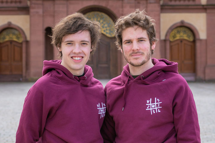 Zwei Studierende vor dem Mannheimer Schloss. Sie tragen beide kurze, braune Haare und einen Hoodie der Fachschaft VWL.