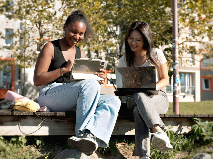 Zwei Studentinnen sitzen bei gutem Wetter mit Laptops auf einer Bank.