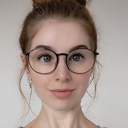 Portrait of Cora Beck. She is wearing a gray hoodie and large round glasses. She has brown eyes and her brown hair is tied up in a bun.