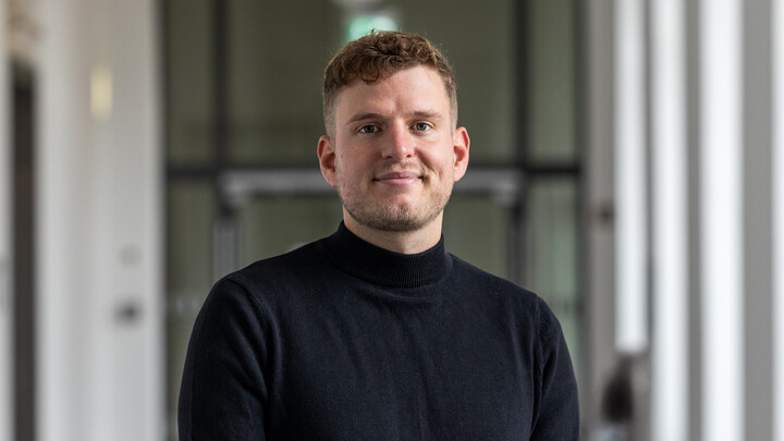 A portrait of Tristan Muno. He is wearing a black sweater and standing in a hallway at the university.