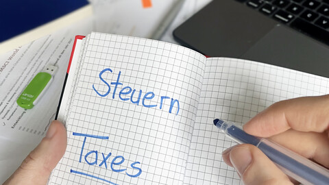 Eine Hand hält ein Notizbuch, in dem Steuern/Taxes eingetragen ist.