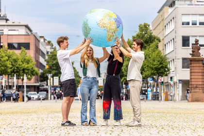 Vier Studierende stehen auf dem Ehrenhof und halten eine aufgeblasene Weltkugel in die Höhe. Im Hintergrund sind Gebäude aus der Stadt zu sehen.