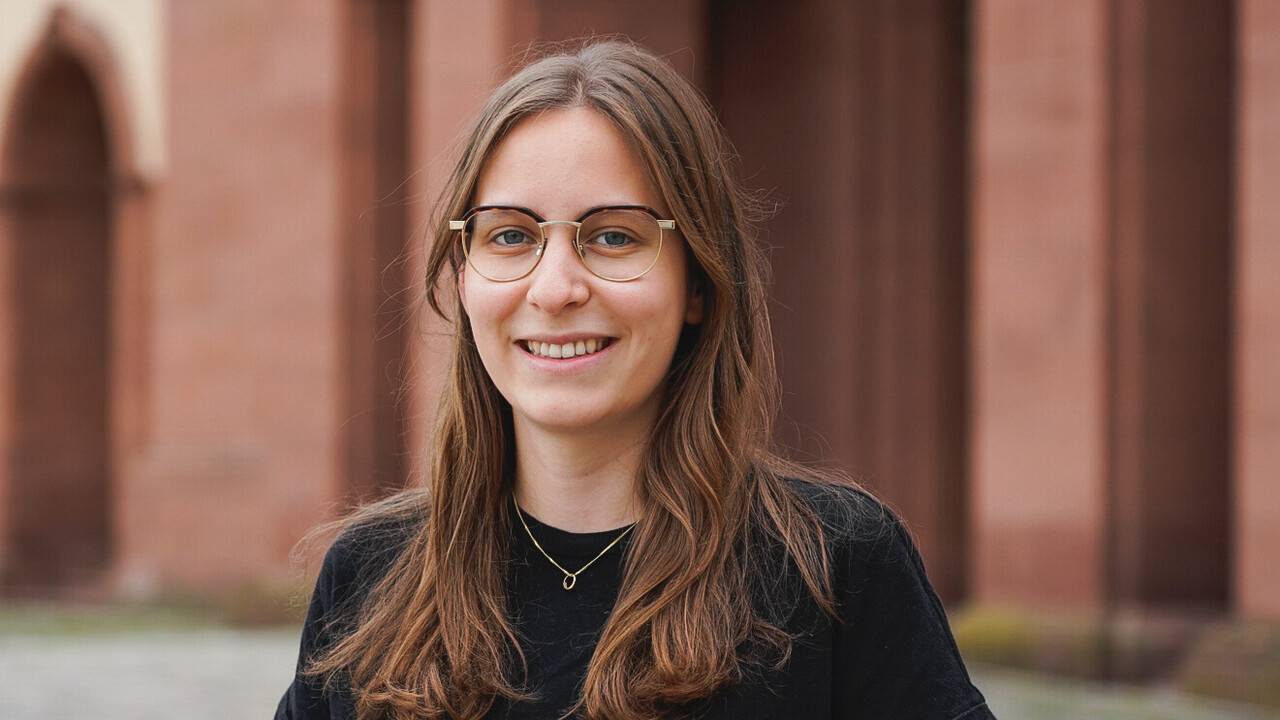 Marlene Fritz hat braune lange Haare. Sie trägt eine goldumrandete Brille und ein schwarzes T-Shirt.