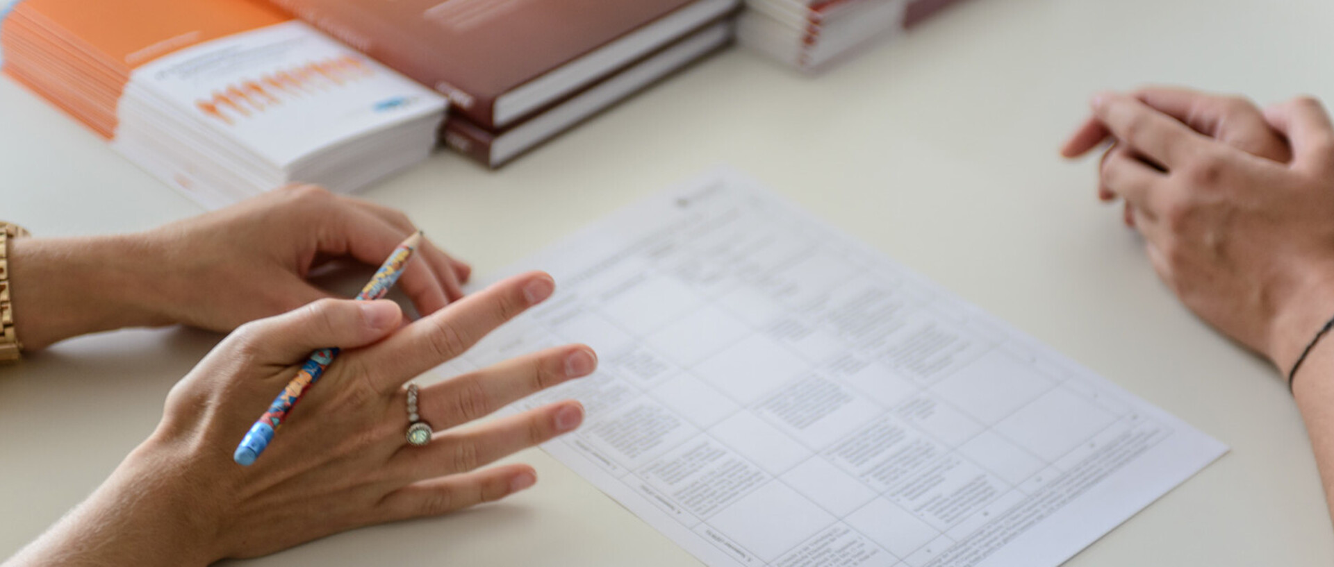 Zwei Personen sitzen an einem Tisch. Weißes Blatt Papier auf dem Tisch. Frau hat Stift in der Hand.