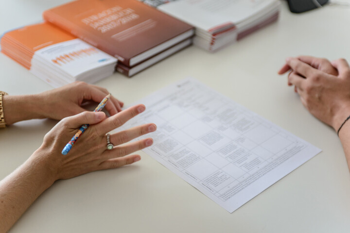 Zwei Personen sitzen an einem Tisch. Weißes Blatt Papier auf dem Tisch. Frau hat Stift in der Hand. 