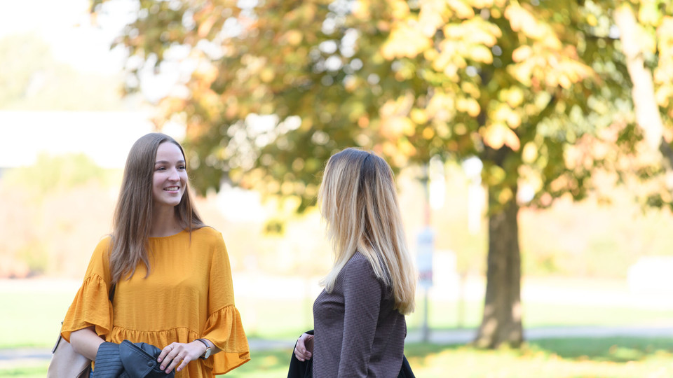 Campusleben Zwei Studierende lächeln. Im Hintergrund ist ein großer Baum.