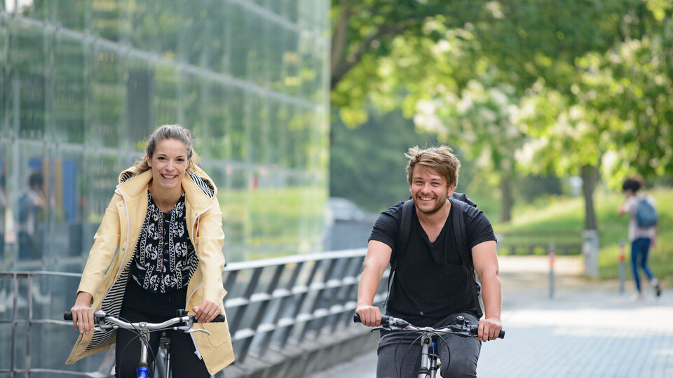 Zwei junge Menschen fahren auf dem Fahrrad an einem Uni-Gebäude vorbei.