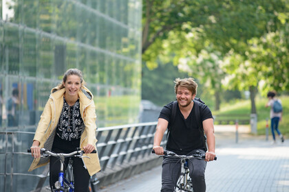 Zwei junge Menschen fahren auf dem Fahrrad an einem Uni-Gebäude vorbei.