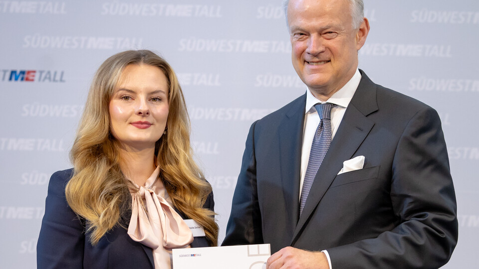 Frau Zercher und Herr Krause stehen vor einer Wand. Sie halten zusammen eine Urkunde und lächeln in die Kamera.