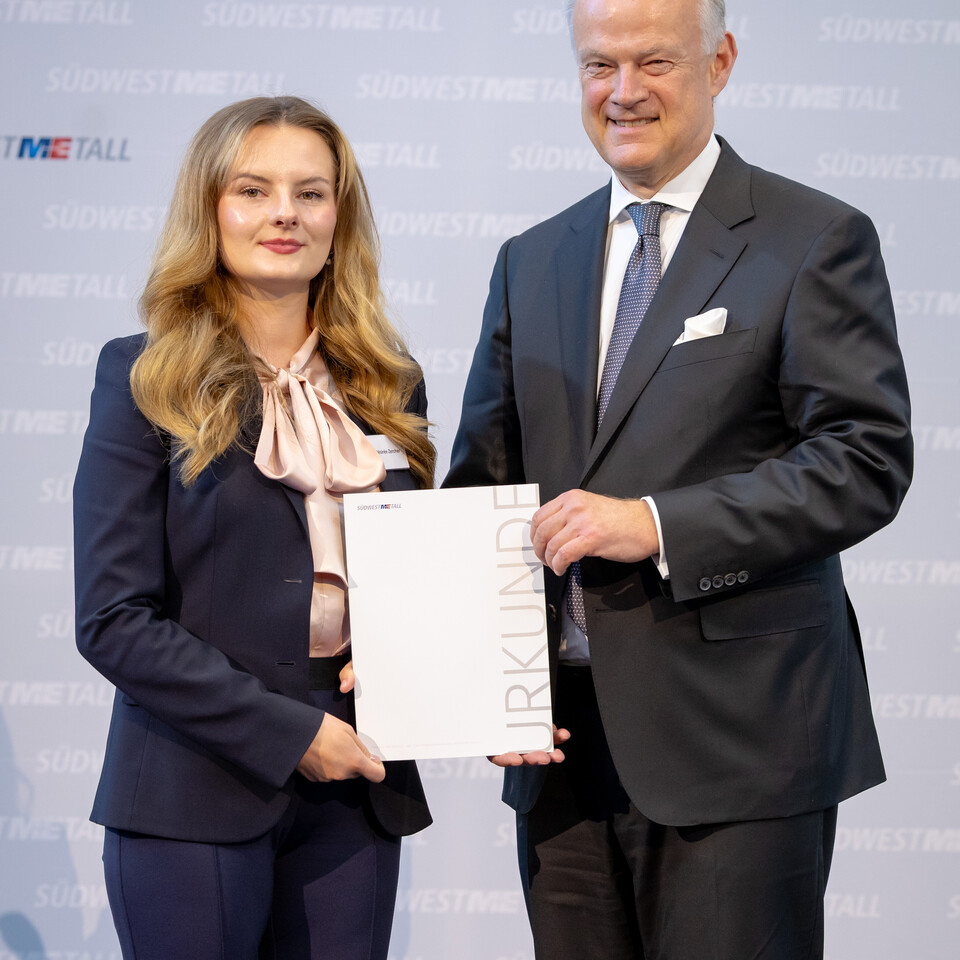 Frau Zercher und Herr Krause stehen vor einer Wand. Sie halten zusammen eine Urkunde und lächeln in die Kamera.