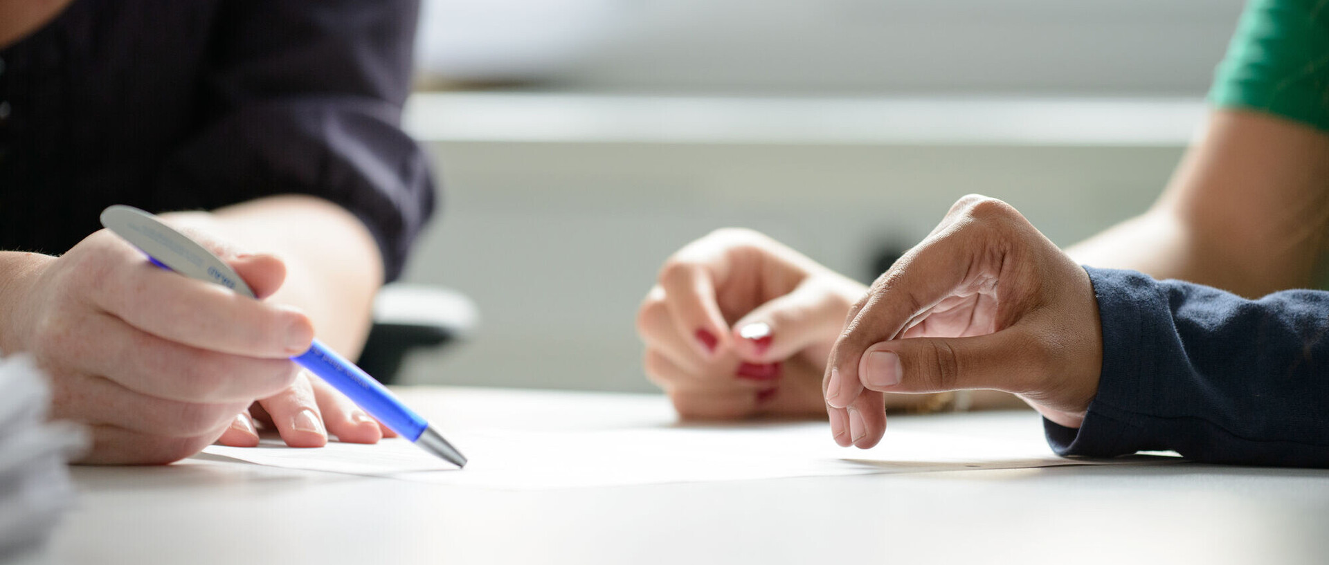 Zwei Studierende werden beraten, dabei sind die Hände und der Stift der Mitarbeiterin, der auf ein Papier zeigt, im Fokus.