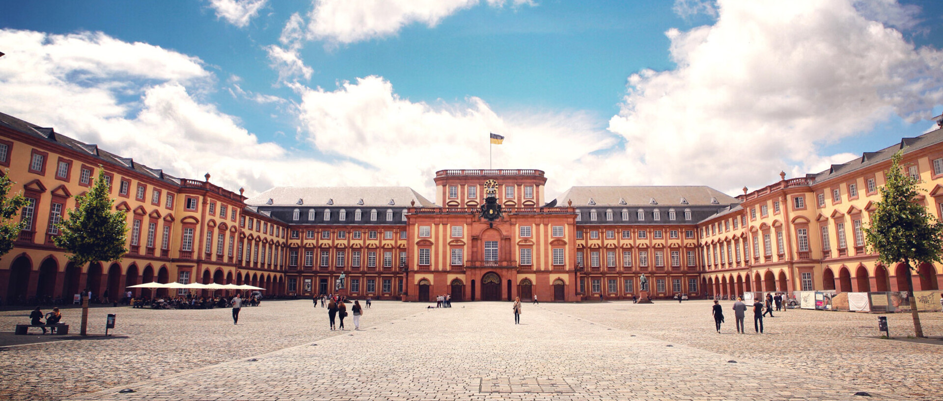 Das Barock-Schloss der Universität Mannheim steht unter blauem Himmel. Es ist von unzähligen Fenstern, rotem Sandstein und einer gelben Fassade geprägt.