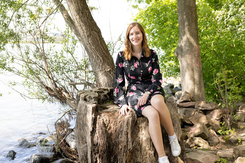 A student in a black dress and pink roses sits on a tree stump by the water.