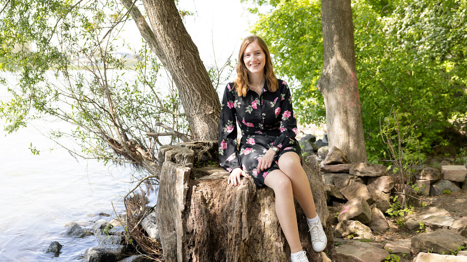 Eine Studentin mit schwarzem Kleid und pinken Rosen sitzt auf einem Baumstumpf am Wasser.