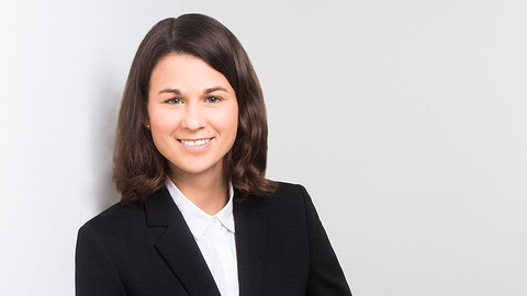 Leah Zimmerer. She is wearing a black suit and is standing in front of white wall.