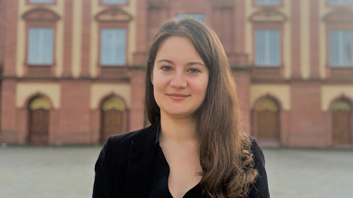 Müge Cakan. She is wearing a black blouse and is standing in front of the castle on the Ehrenhof.