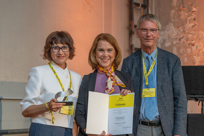 Professorin Kogan steht mit Ministerin Olschowski und mit Hans-Georg Kräusslich auf der Bühne und hält ihre Auszeichnung in der Hand.