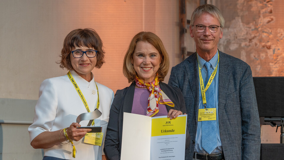 Professorin Kogan steht mit Ministerin Olschowski und mit Hans-Georg Kräusslich auf der Bühne und hält ihre Auszeichnung in der Hand.