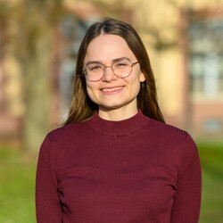 Paula Osburg hat lange braune Haare, trägt eine Brille und ein dunkelrotes Oberteil. Sie lächelt in die Kamera, im Hintergrund ist die Grünanlage vor dem Schloss zu sehen. 