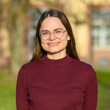 Paula Osburg hat lange braune Haare, trägt eine Brille und ein dunkelrotes Oberteil. Sie lächelt in die Kamera, im Hintergrund ist die Grünanlage vor dem Schloss zu sehen. 