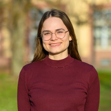 Paula Osburg hat lange braune Haare, trägt eine Brille und ein dunkelrotes Oberteil. Sie lächelt in die Kamera, im Hintergrund ist die Grünanlage vor dem Schloss zu sehen. 