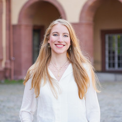 Eine lächelnde Person trägt eine weiße Bluse und steht vor dem Schloss der Uni Mannheim. Die Person heißt Sydney Greiner.