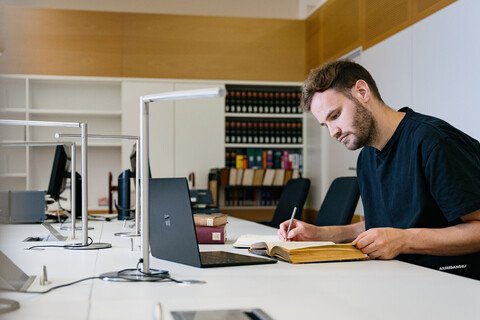 Ein Studierender mit Buch und Laptop in der Bibliothek