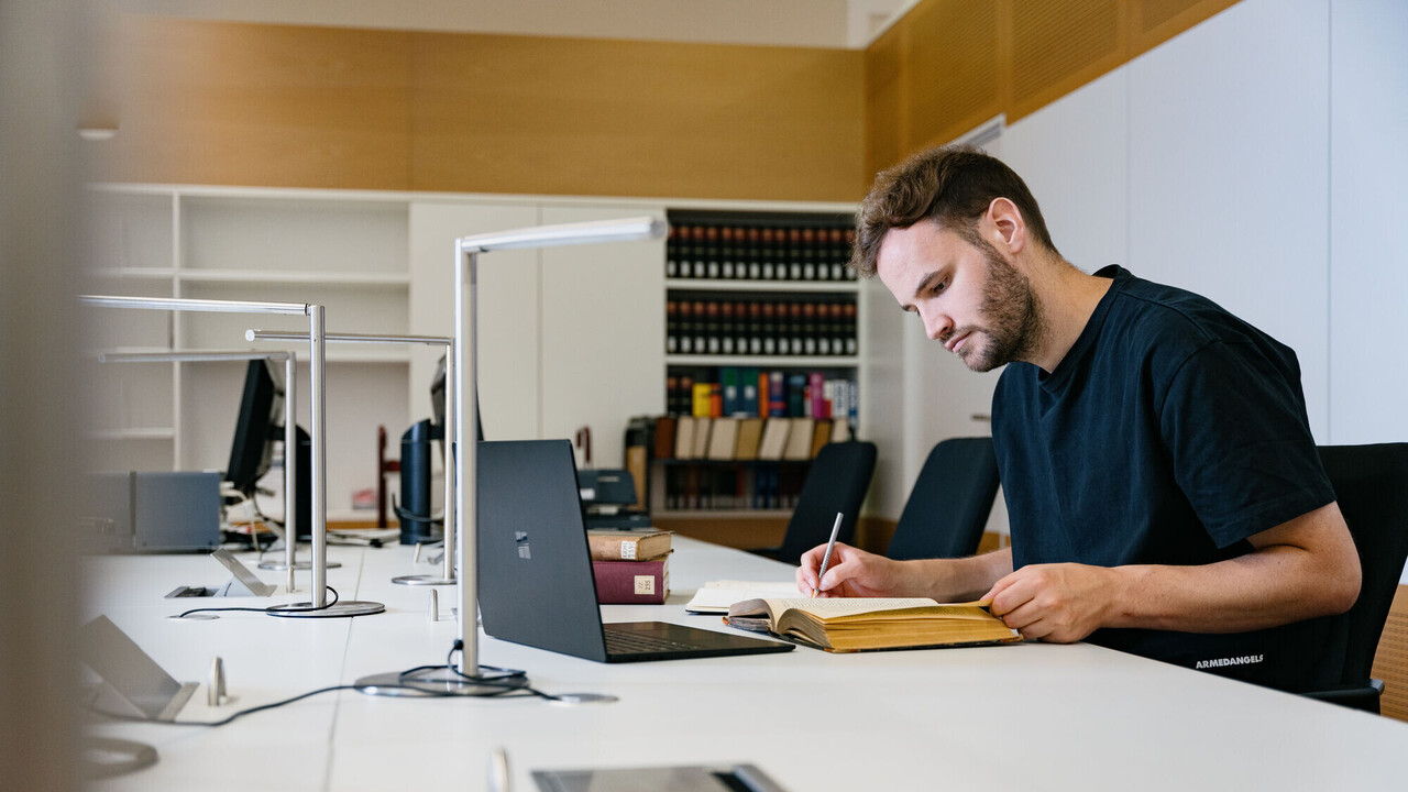 Ein Studierender mit Buch und Laptop in der Bibliothek