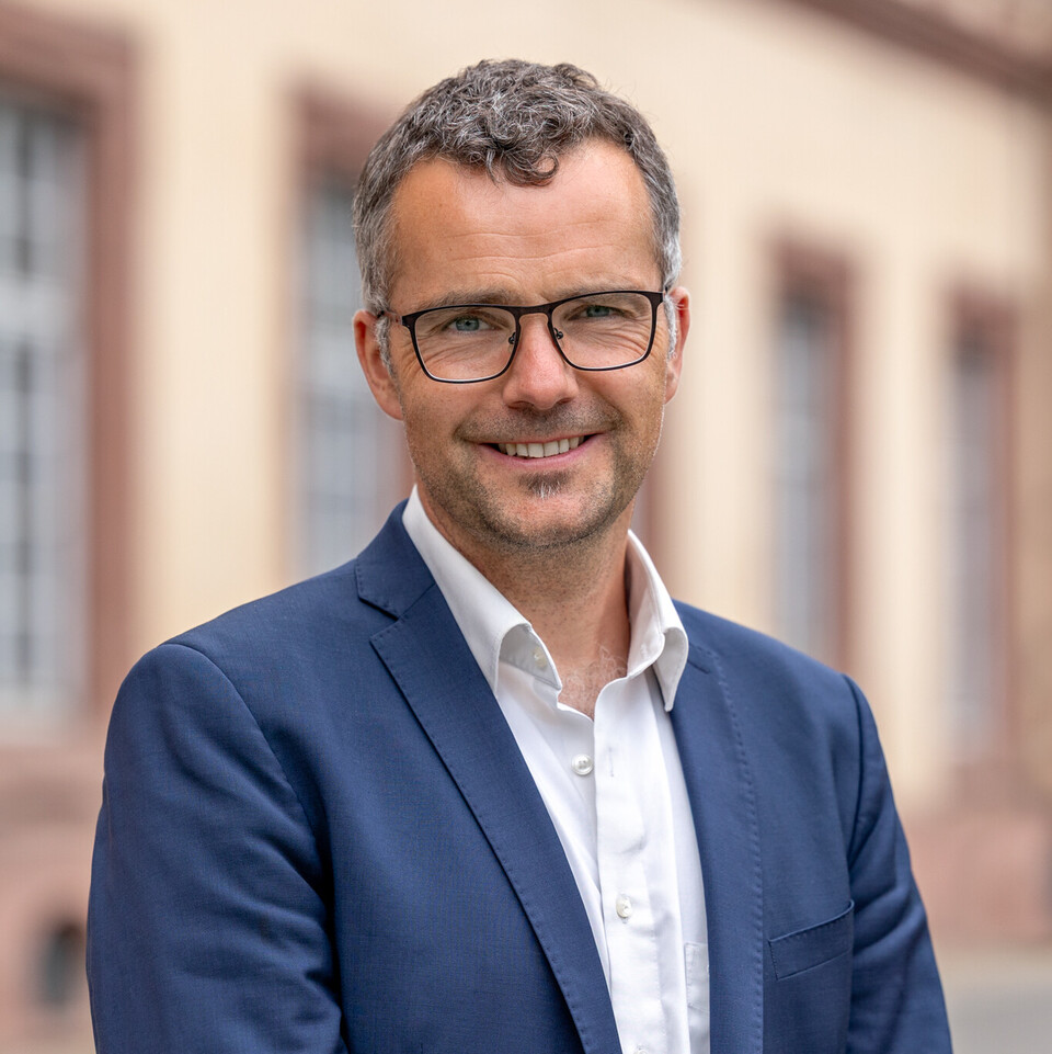 Prof. Debus steht vor dem Schloss und lächelt. Er trägt blauen Anzug und ein weißes Hemd. 