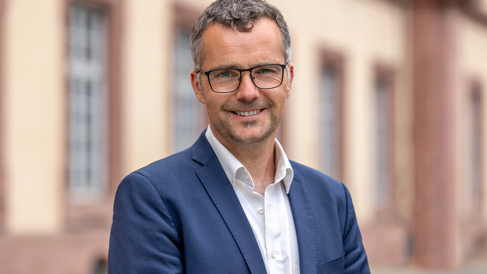 Prof. Debus steht vor dem Schloss und lächelt. Er trägt blauen Anzug und ein weißes Hemd. 