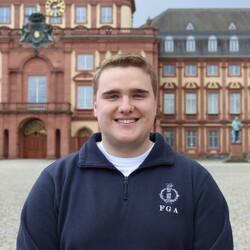 Philipp, mit kurzen blonden Haaren trägt einen dunkelblauen Fleecepullover der Fachschaft und lächelt vor dem Mannheimer Schloss in die Kamera