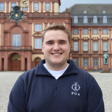 Philipp, mit kurzen blonden Haaren trägt einen dunkelblauen Fleecepullover der Fachschaft und lächelt vor dem Mannheimer Schloss in die Kamera