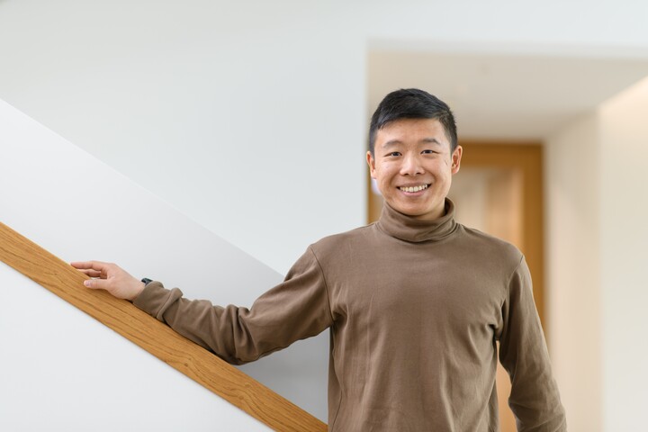 Jianming Cui aus Kanton. Er trägt einen braunen Rollkragenpullover und legt seinen rechten Arm lässig auf ein Treppengeländer. Link: myUniMa story Mai 2023