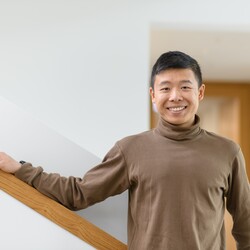 Jianming Cui stands smiling in a hallway. He is wearing a light brown turtleneck sweater.