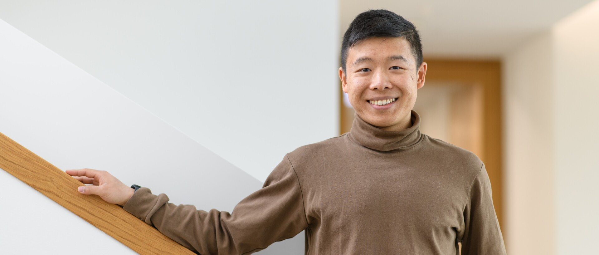 Jianming Cui stands smiling in a hallway. He is wearing a light brown turtleneck sweater.