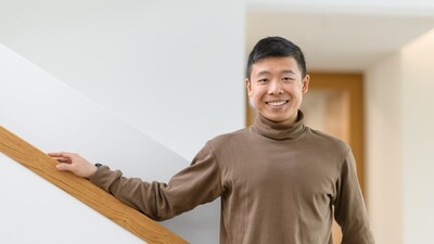 Jianming Cui stands smiling in a hallway. He is wearing a light brown turtleneck sweater.