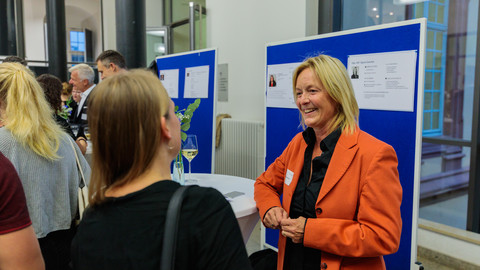 An einem Messestand steht eine Person mit orange-farbenem Blazer und spricht mit einer weiteren Person. Im Hintergrund sind weitere Messestände aufgebaut, um welche sich Personen versammelt haben.