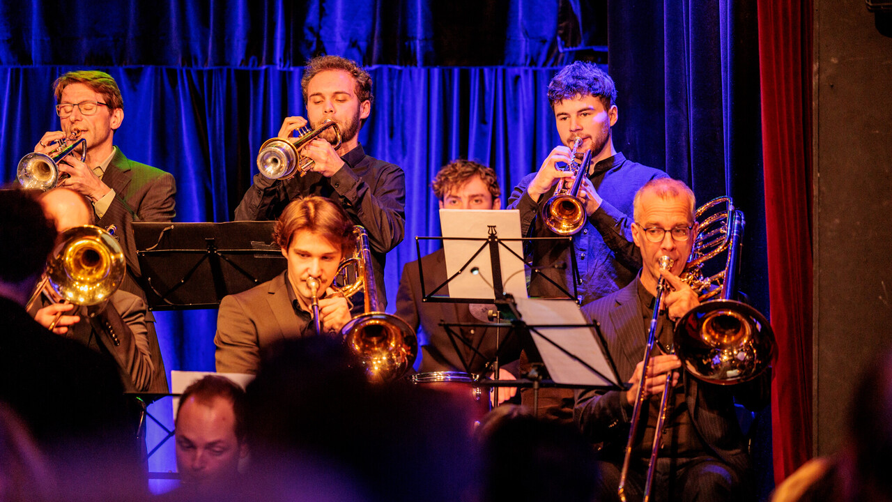 Acht Musiker einer Bigband spielen verschiedene Blasinstrumente auf einer Bühne.