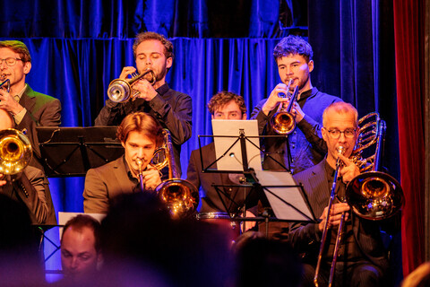 Acht Musiker einer Bigband spielen verschiedene Blasinstrumente auf einer Bühne.