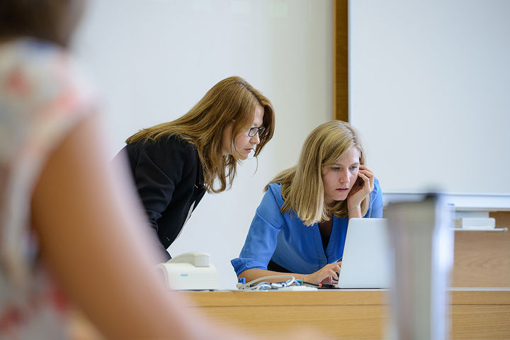 Zwei Frauen schauen zusammen auf den Display eines Laptops. Sie stehen vorne an der Tafeln.