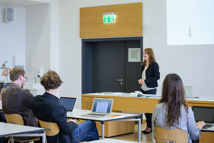 Eine Dozentin steht lachend im Seminar vor drei Personen, die in der vorderen Reihe sitzen.