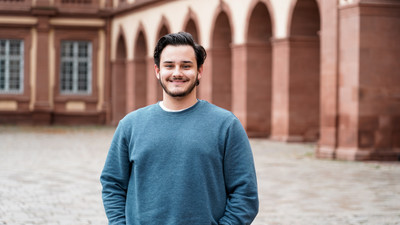 Eine Person trägt einen blauen Pullover sowie eine dunkelblaue Jeans und steht vor dem Schloss der Uni Mannheim. Die Person heißt Felipe Gomes dos Reis Justininano.