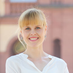 Lea Tochtermann hat ihre blonden Haare zu einem Zopf gebunden und hat einen Pony im Gesicht. Sie trägt eine weiße Bluse und lächelt in die Kamera.